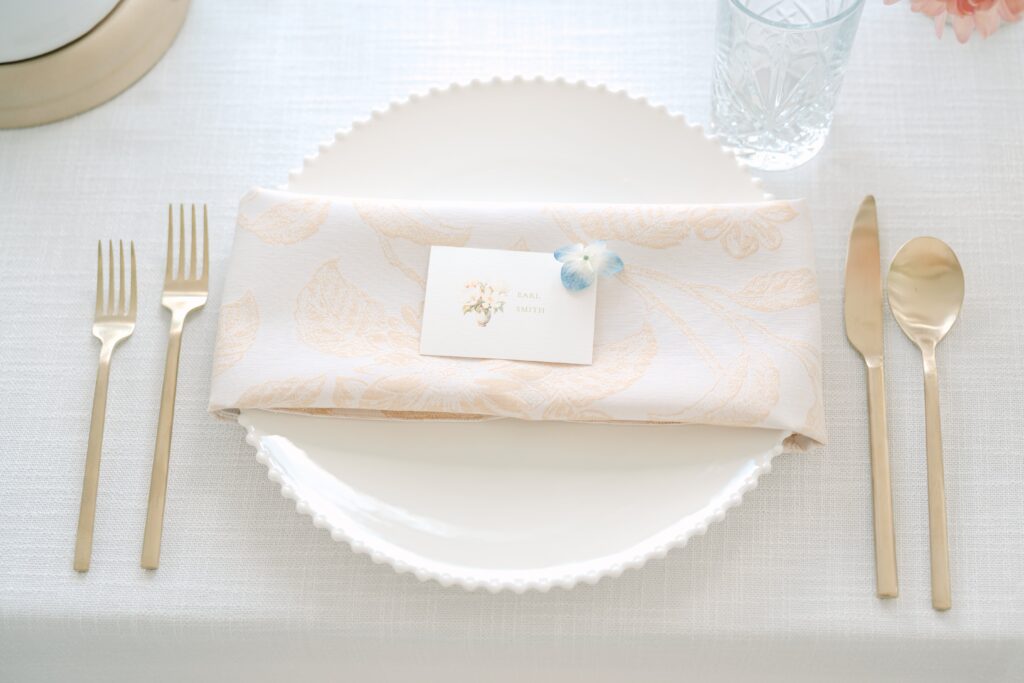 Delicate wedding place card with floral watercolor art sits atop a white and champagne colored napkin