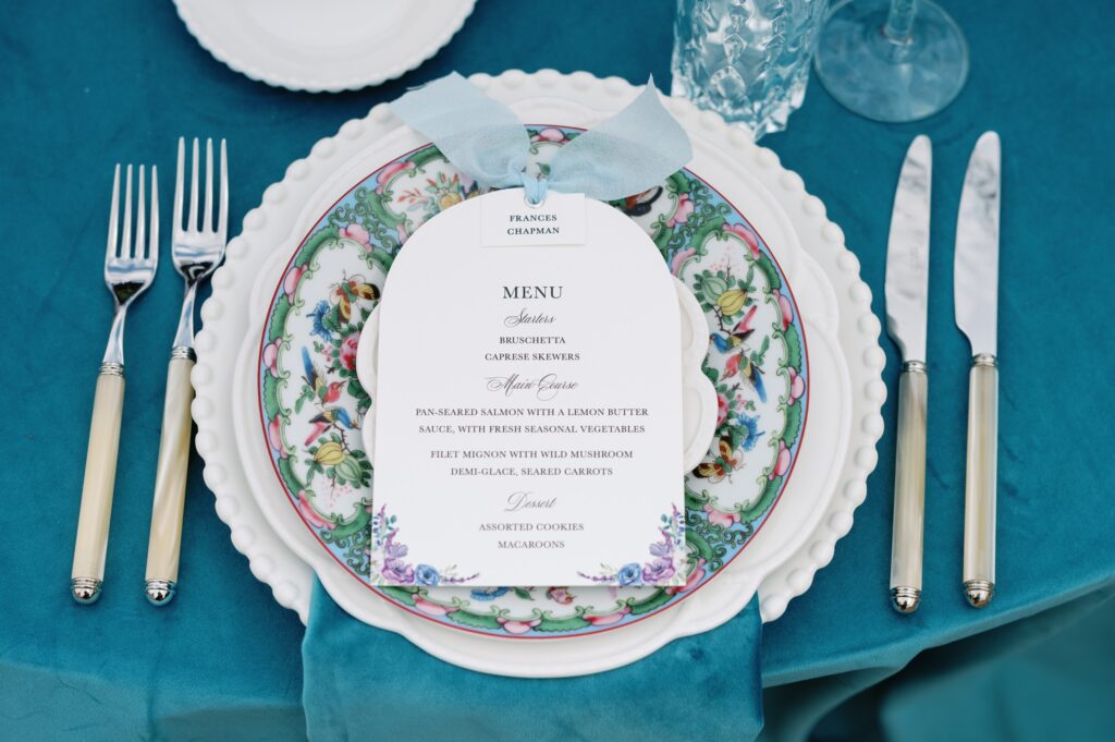 Die-cut wedding menu card with a blue bow and elegant watercolor florals sitting on vintage floral plates