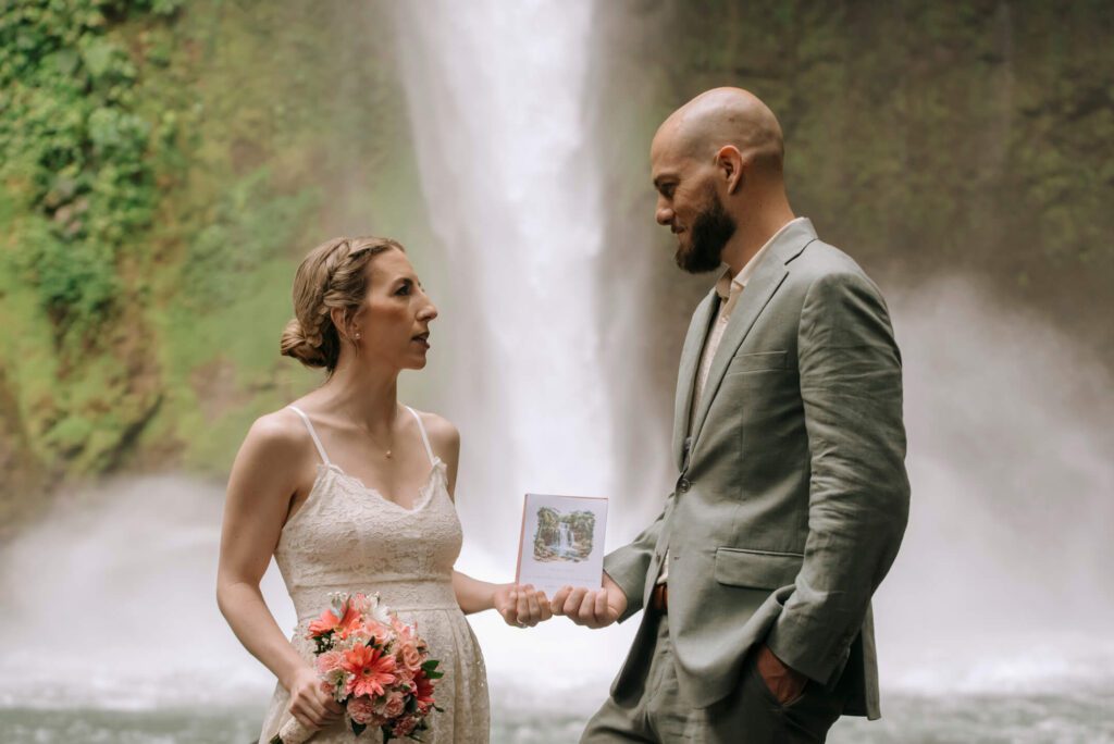 Bride and groom holding custom elopement announcement card in front of La Fortuna Waterfall