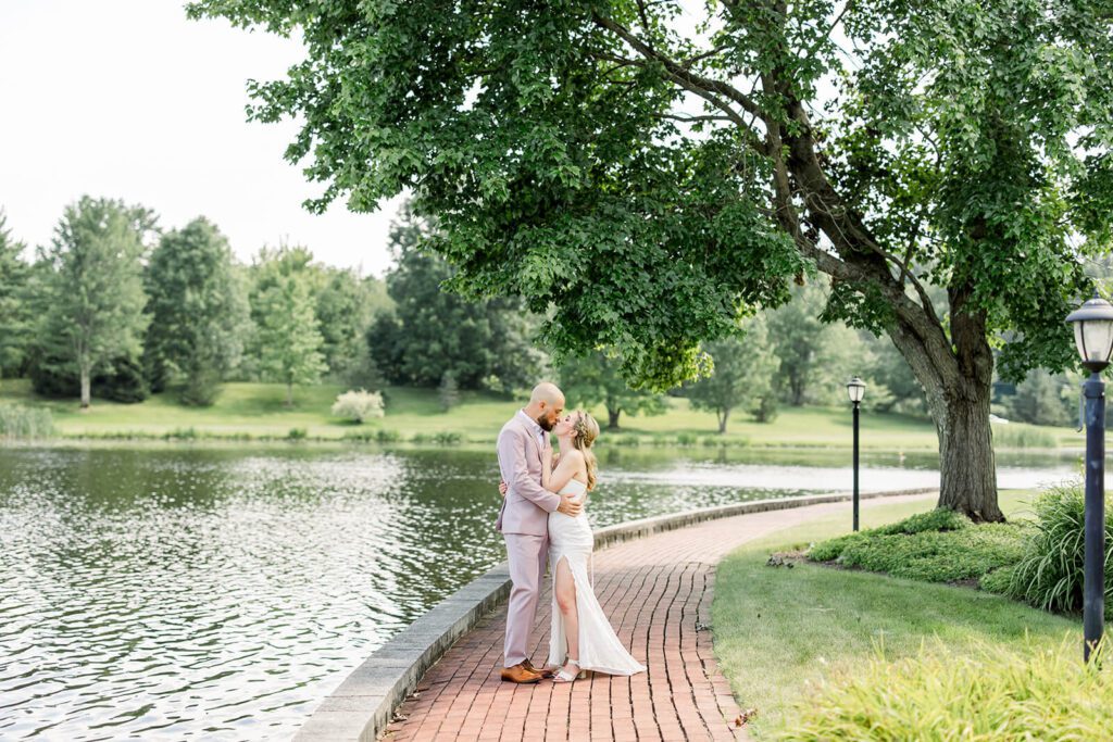 Bride and groom kiss outdoors by a pond surrounded by lush greenery on their wedding day