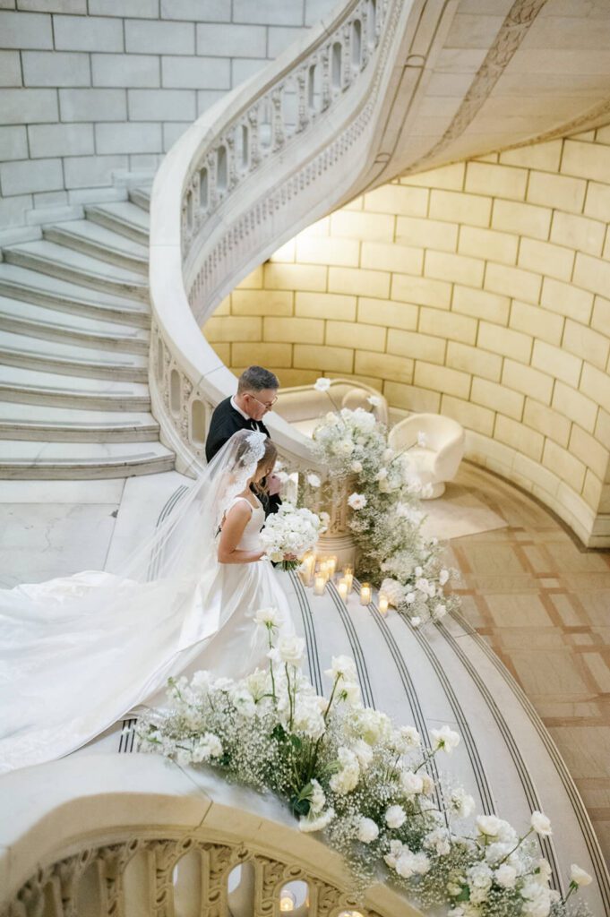 Bride escorted by her father down an elegant marble staircase in The Old Courthouse