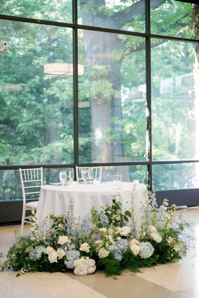 Elegant white and blue floral sweetheart table in front of floor to ceiling windows showing the forest