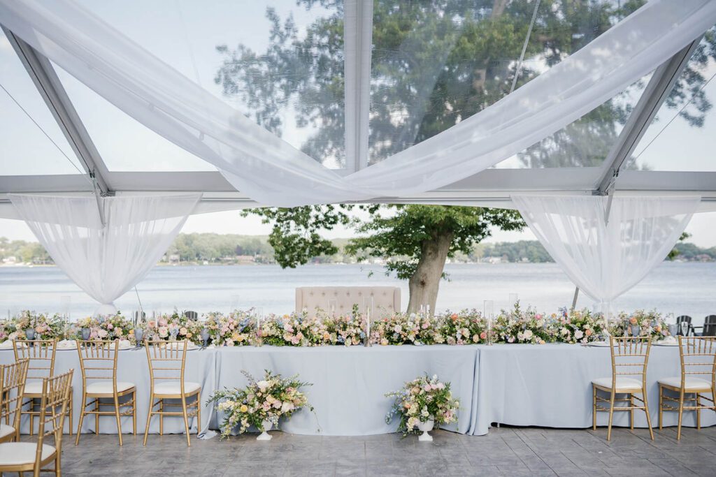 Elegant wedding reception head table by the water with greenery and white draping