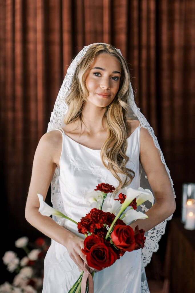 Portrait of bride wearing lacy cathedral veil and holding red and white bouquet