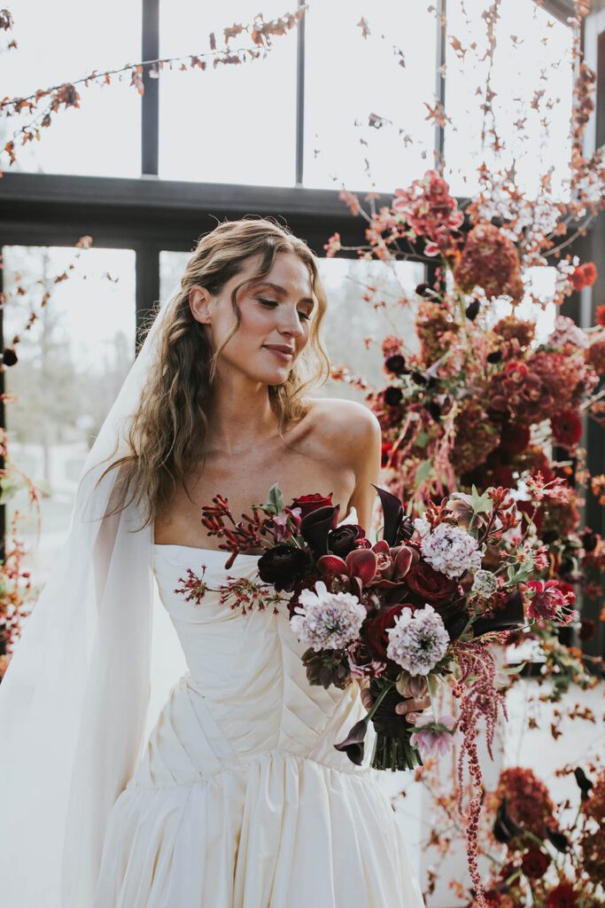 Bride wearing drop waist corseted wedding dress and holding a romantic bouquet in shades of red