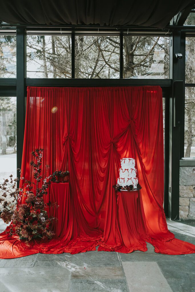 Dramatic red drapery backdrop with two pedestals. The one on the left contains drinks and the one on the right contains a vintage cake with red bows