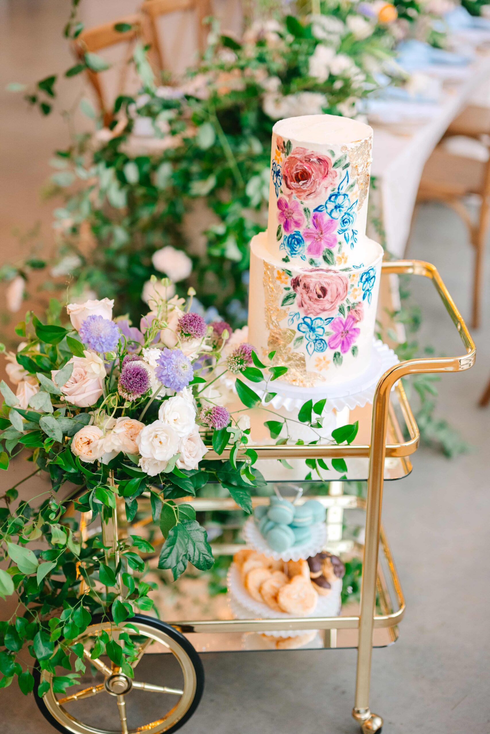 Two-tiered wedding cake with purple and blue flowers sitting on a gold cart