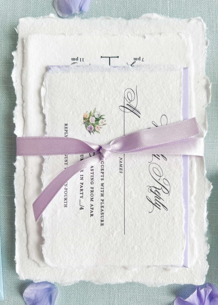 Deckled edge wedding invitation suite cards stacked and tied with a purple ribbon